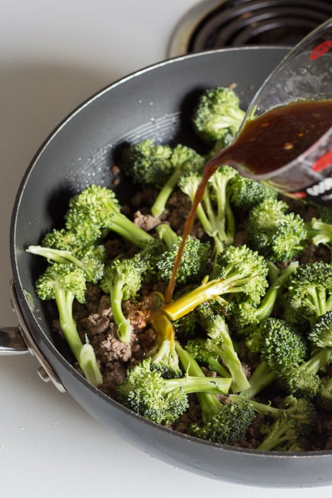 Stir fry sauce being poured over the ground beef and broccoli in a large black pan on the stove.