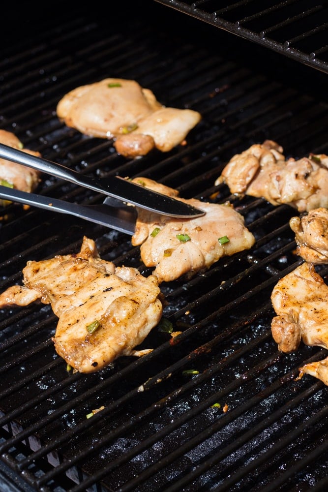 A tong place a boneless skinless teriyaki chicken thigh on a Traeger grill grate.