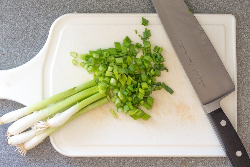 Top down shot of chopped green onions on a white cutting board with a large sharp knife next to them.