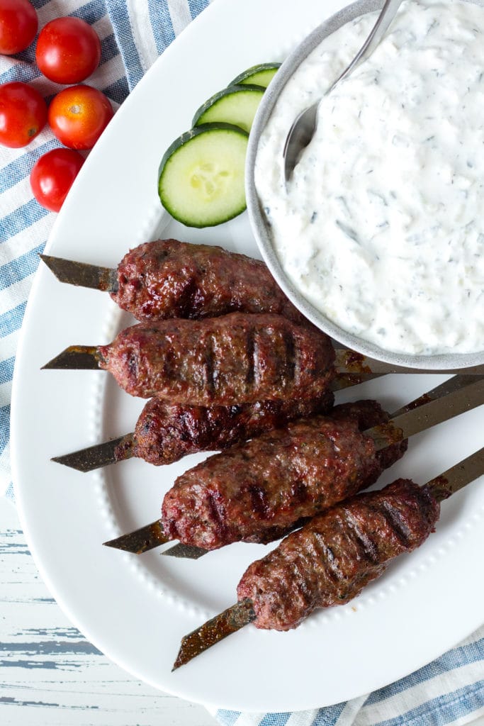 Top down view of lamb skewers on a white platter with cucumber and cherry tomatoes on it, and a small gray bowl with tzatziki sauce in it.