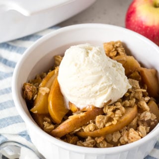 small white cup with apple crisp topped with ice cream on top of a striped blue napkin with apples in the background