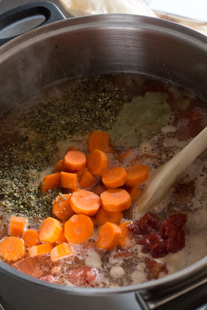 A large pot with soup ingredients in it, including herbs, sliced carrots, tomato paste, and a bay leaf. A wooden spoon rests in the pot as well.