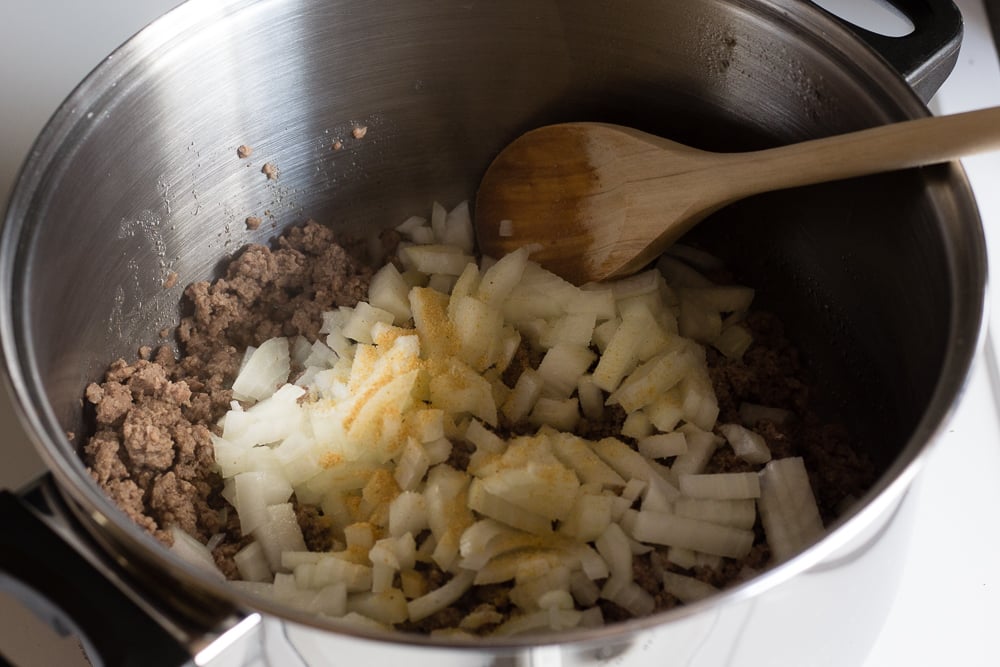Ground beef and onions in a large silver pot with spices. A wooden spoon rests in the pot as well.