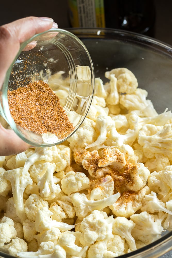A hand pouring spices from a small glass bowl into a larger bowl with cauliflower pieces in it.