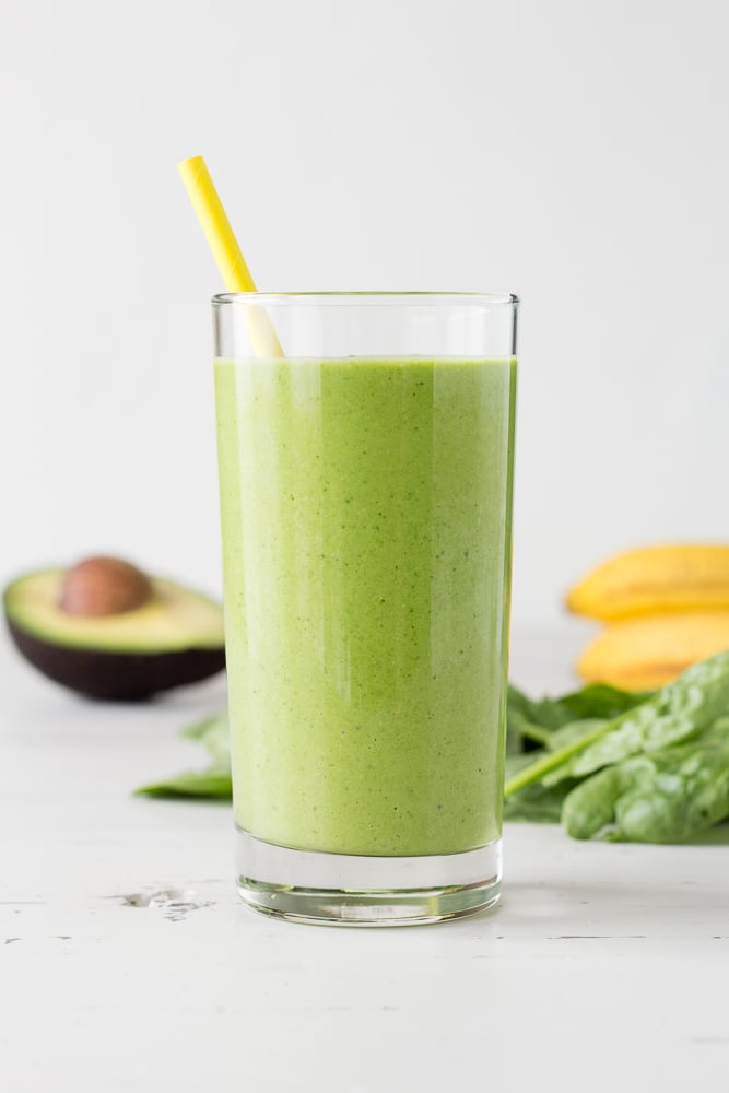 Straight on shot of a green smoothie in a clear glass with a yellow paper straw sticking out of it. Half an avocado, some spinach, and bananas are in the background.
