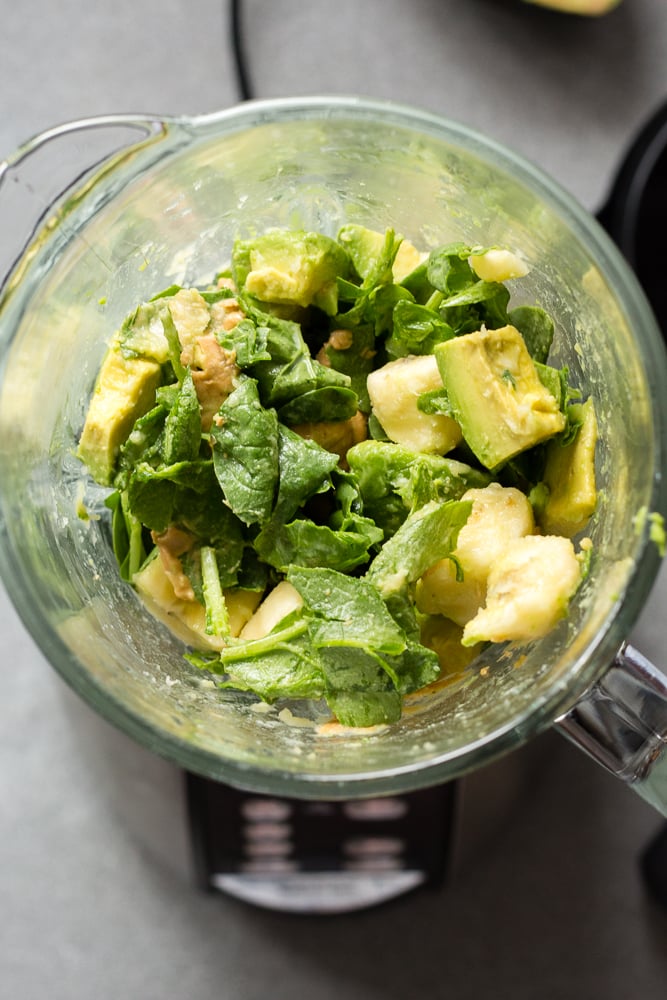 Chopped up smoothie ingredients in a blender, including spinach, avocado, banana, and nut butter.
