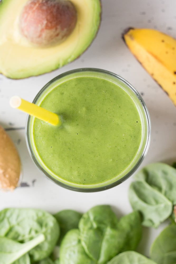 Top down shot of a green smoothie in a glass with a yellow straw, surrounded by avocado, banana, spinach, and nut butter.