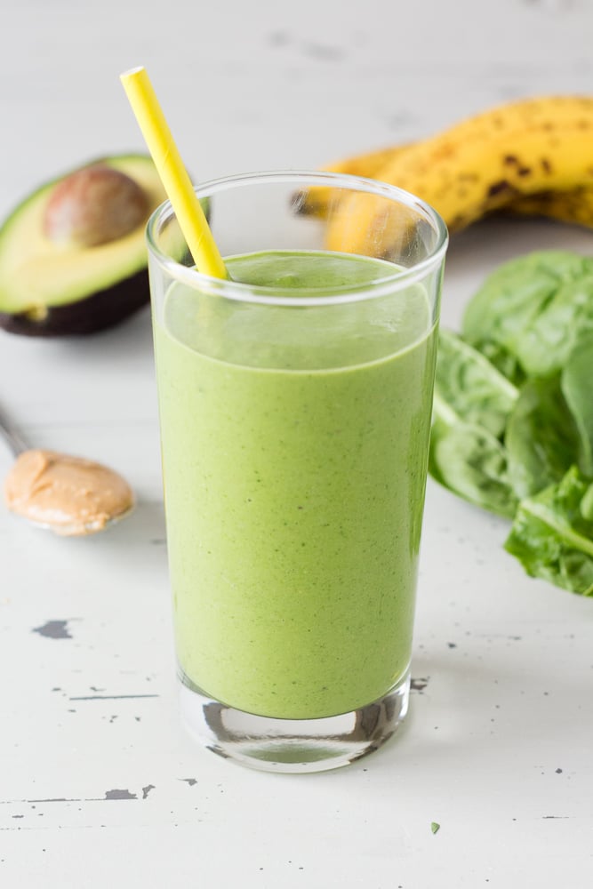 Close up of a green smoothie in a glass with a yellow straw. Half an avocado, some baby spinach, bananas, and a spoonful of nut butter are in the background.