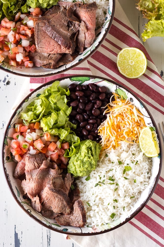 Top down shot of two bowls with white rice, shredded cheese, black beans, lettuce, pico de gallo, sliced steak, and guacamole in them. Cut lime and a red and white striped towel lay next to the bowls.