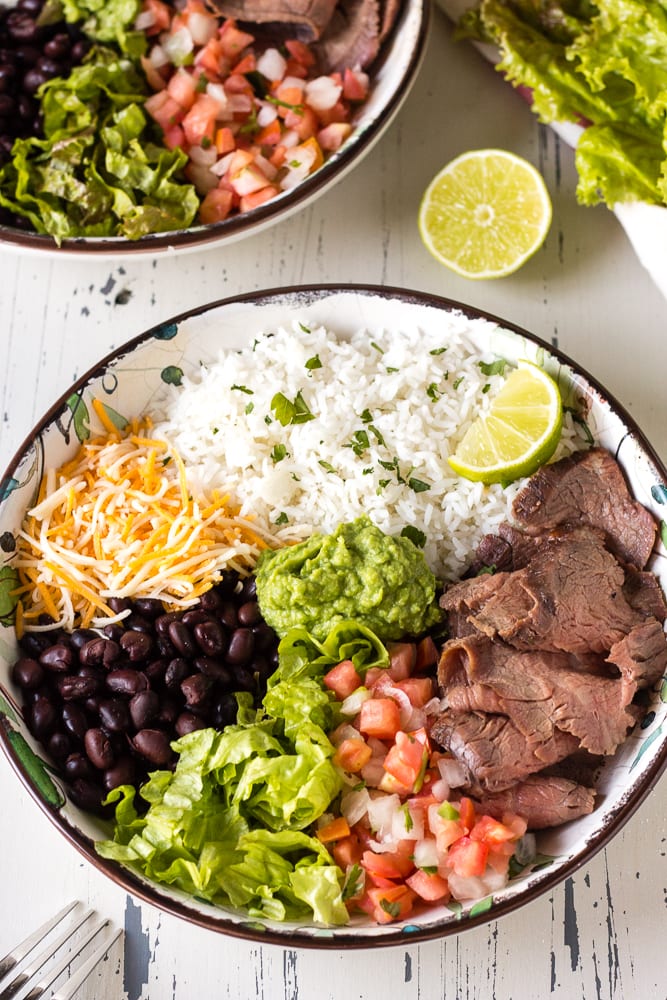 Top down shot of shredded cheese, black beans, chopped lettuce, pico de gallo, white rice, and sliced flank steak in a bowl with guacamole in the middle and a slice of lime on the side.