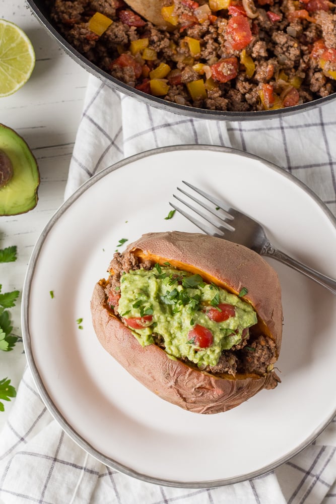Top down shot of beef stuffed sweet potato with beef and guacamole on a white plate with a fork, next to a skillet with ground beef in it. Half an avocado and a cut lime are next to the plate.