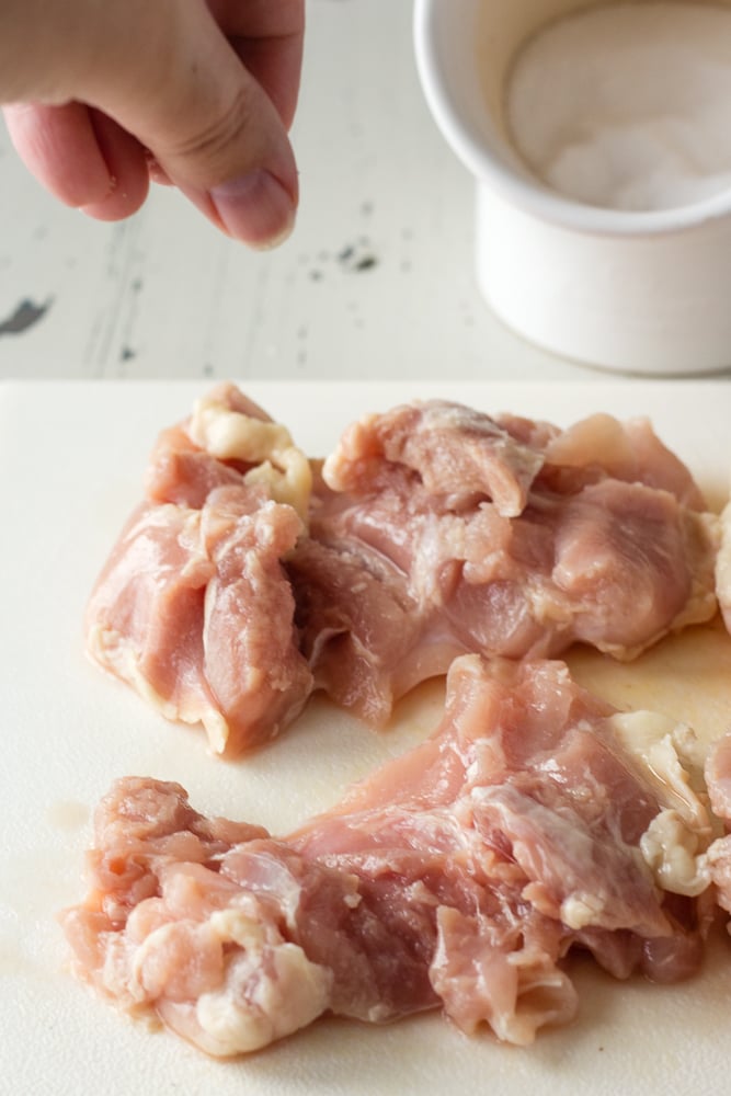 A hand sprinkling salt over boneless skinless chicken thighs on a white cutting board.