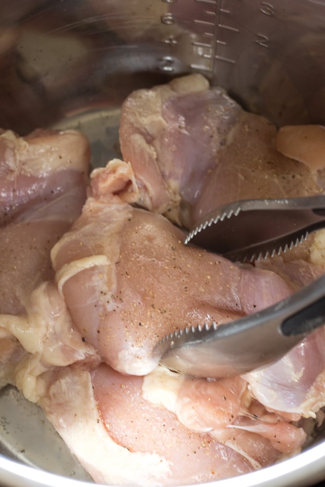 A pair of tongs placing raw chicken thighs in an Instant Pot.