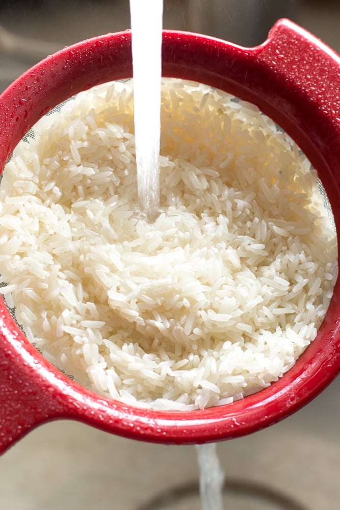 A red handheld colander with white rice in it being rinsed in a sink.