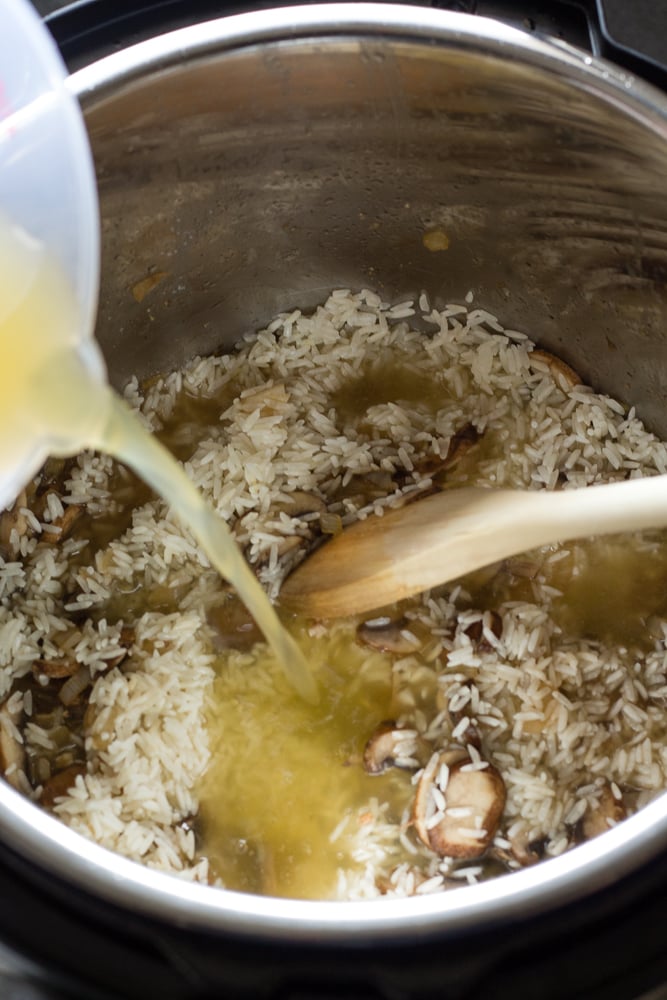 Pouring chicken broth onto white rice, diced onion and sliced mushrooms. A wooden spoon is also in the picture.