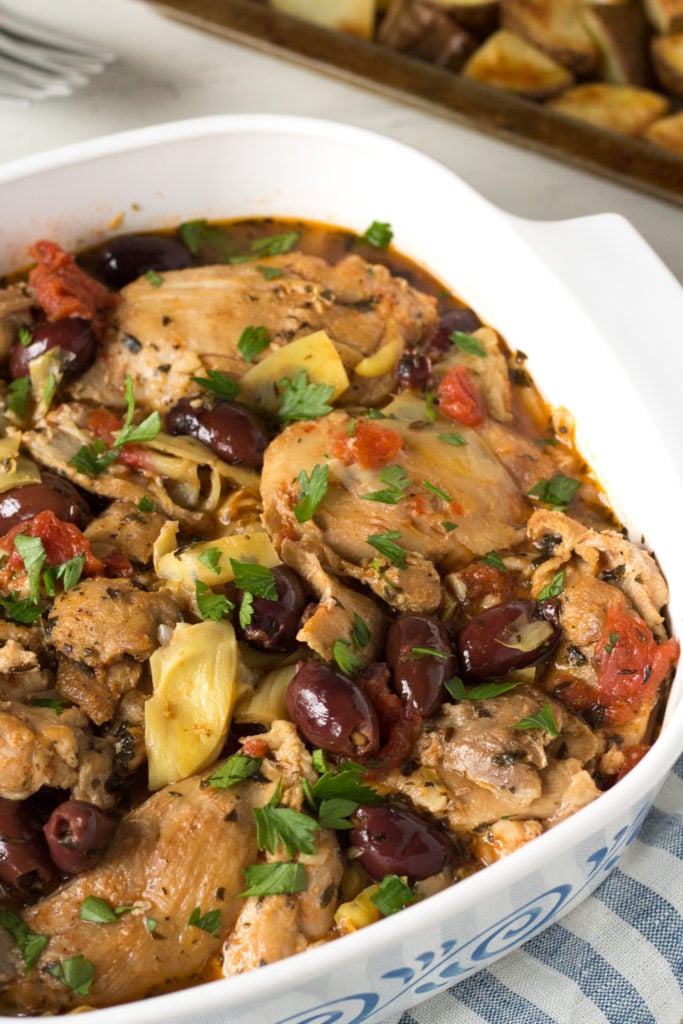 A close up shot of Greek chicken thighs in a white square dish with kalamata olives, artichoke hearts, diced tomatoes, and chopped parsley sprinkled on top.