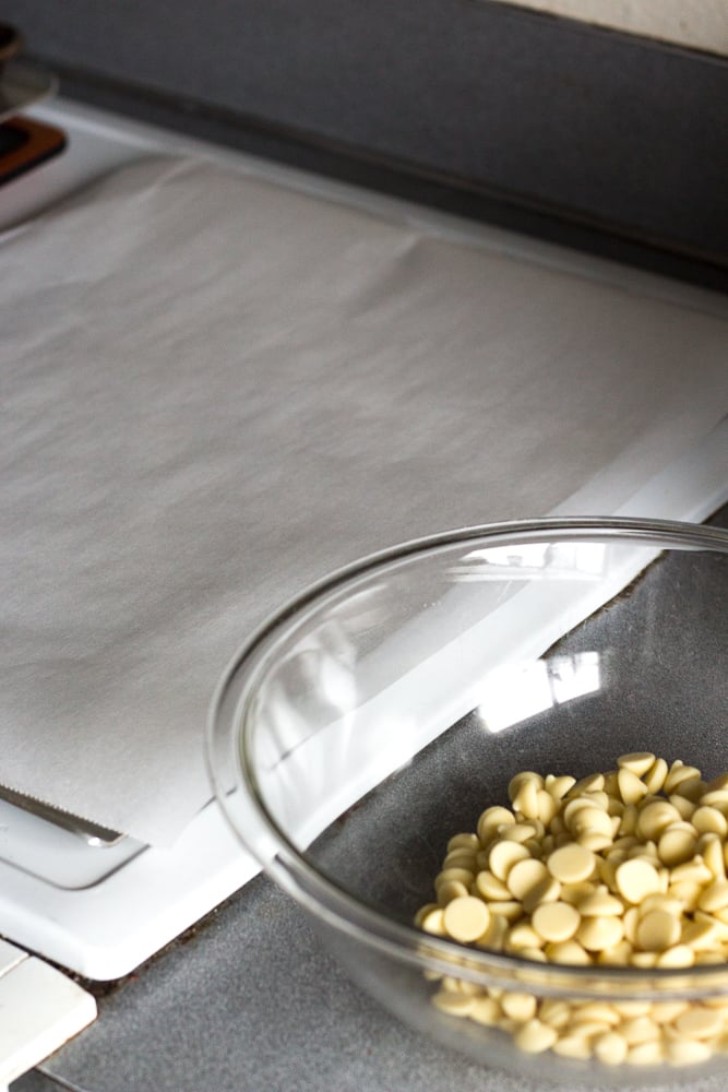 A piece of white parchment on a sheet pan next to a clear bowl with white chocolate chips.