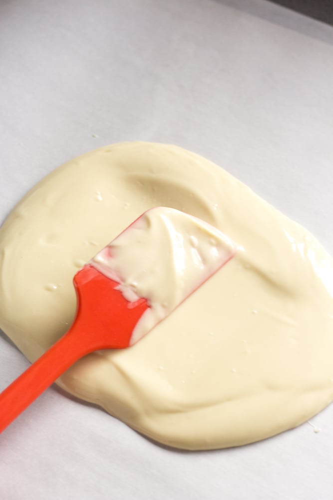 A red spatula spreading white chocolate on a sheet pan lined with parchment paper.