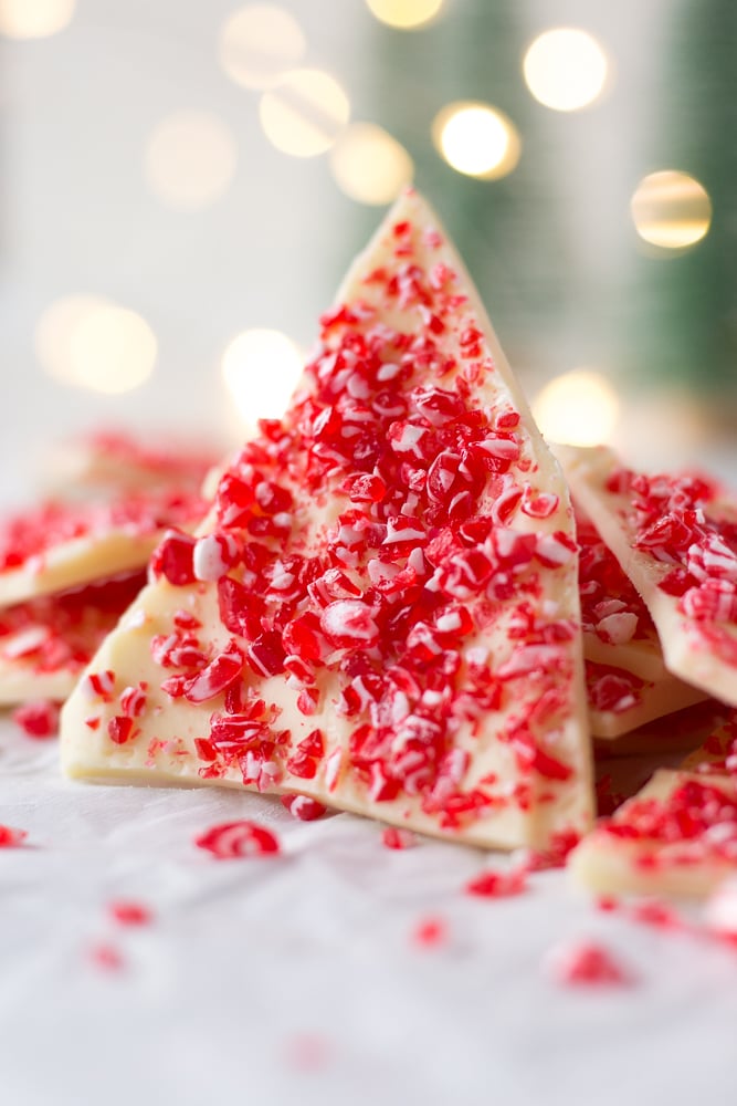 A triangular piece of white chocolate peppermint bark standing up against other pieces of peppermint bark. Twinkly lights are in the background.