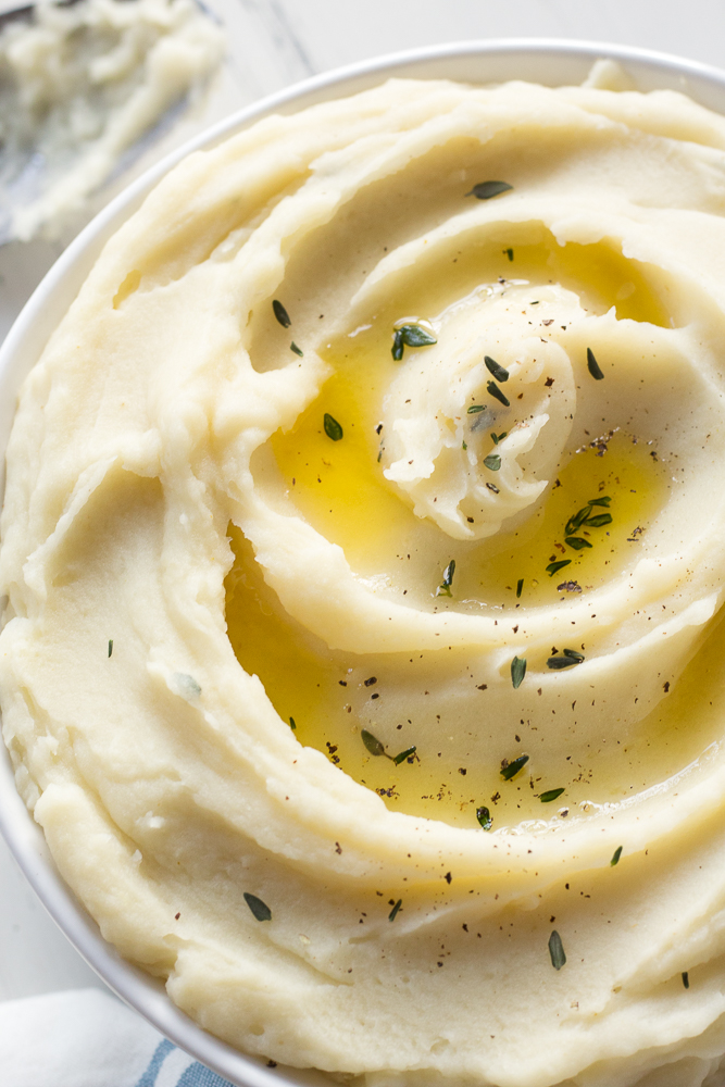 Close up of a big bowl of mashed potatoes drizzled with ghee and sprinkled with herbs and black pepper, with a spoon on the side.