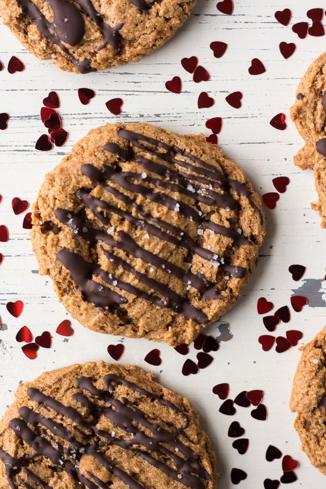 Top down shot of flourless almond butter cookies with chocolate drizzled on top and heart confetti scattered around.