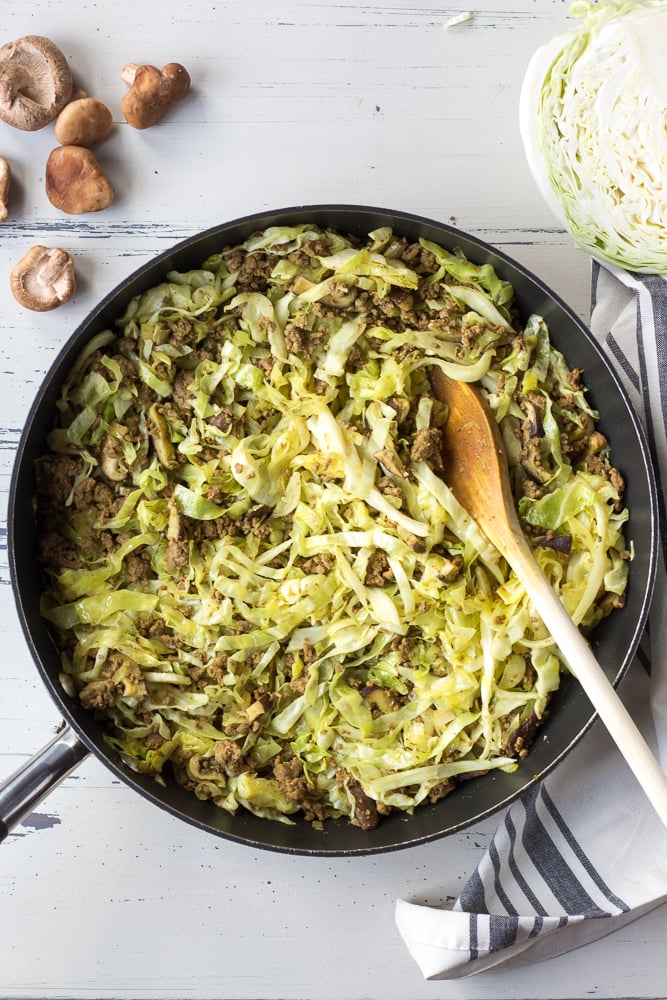 Top down shot of a large black pan with beef and cabbage stir fry in it. A wooden spoon sticks out of it and mushrooms and cabbage are next to it.