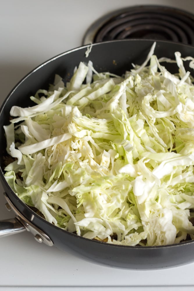 Thinly sliced raw cabbage in a large black pan with beef stir fry on a stove burner.