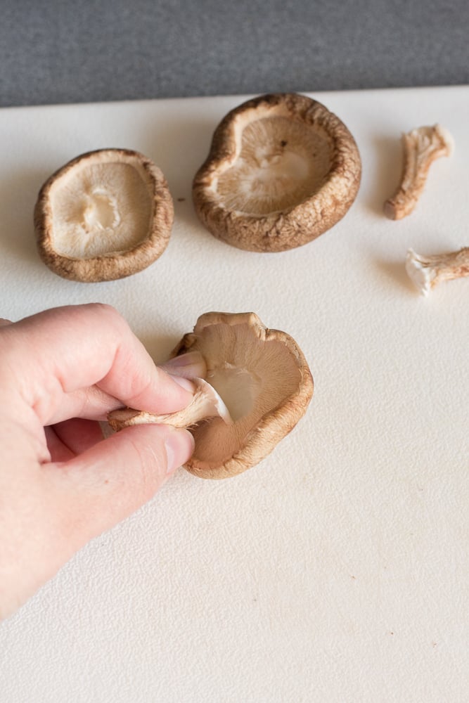 A hand pulling the stem off a shiitake mushroom on a white cutting board.
