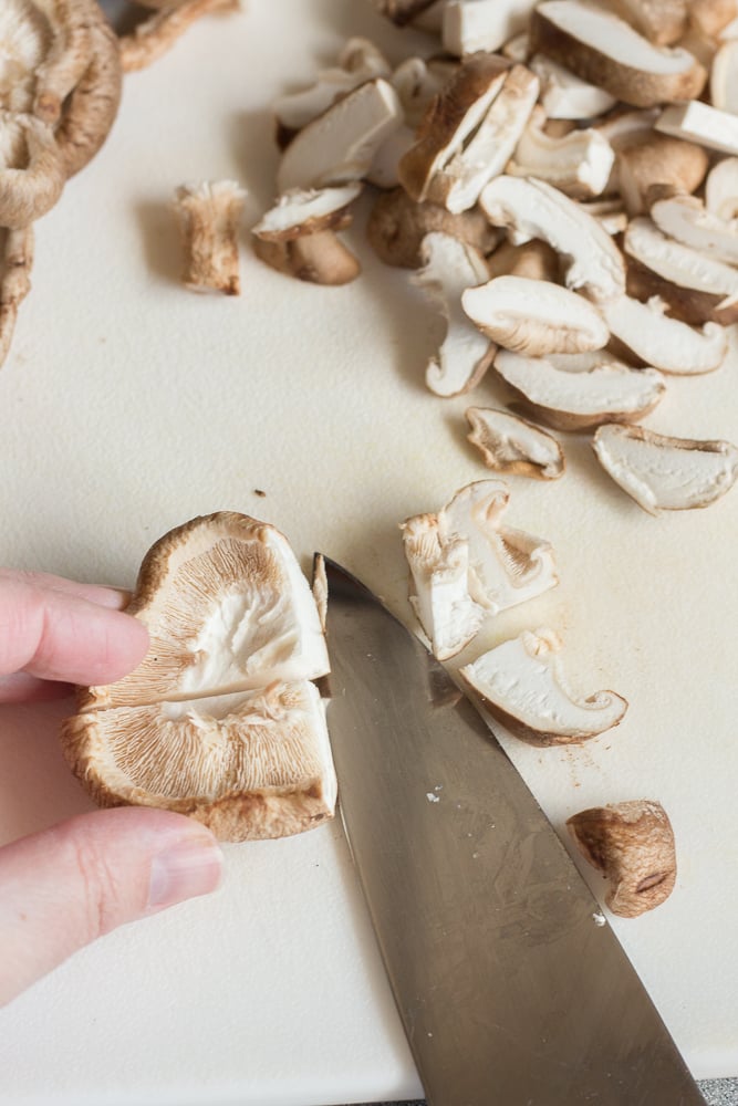 A hand cutting shiitake mushrooms with a knife on a white cutting board.