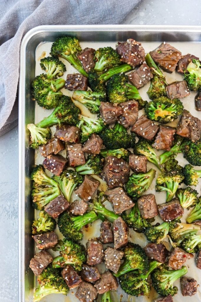 Top down close up of broccoli florets and cubed beef on a sheet pan with sesame seeds on top.