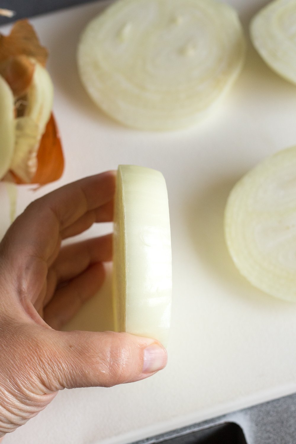 A hand holding up a large slice of an onion on a white cutting board. Other slices of onion lay on the cutting board around it.
