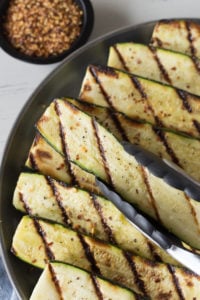 tongs placing grilled zucchini on a gray plate