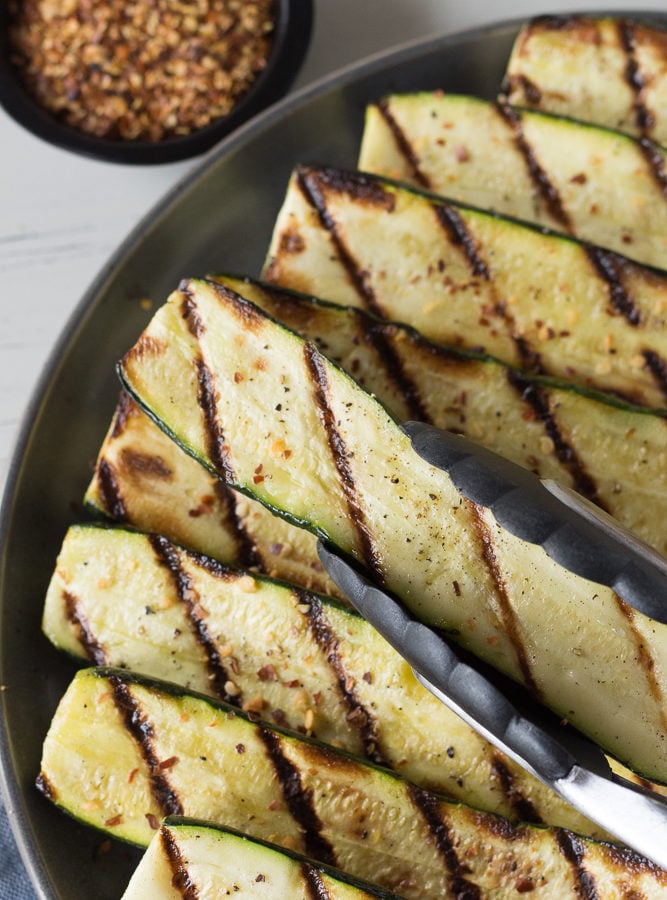 Top down shot of a pair of tongs placing grilled zucchini on a gray plate.