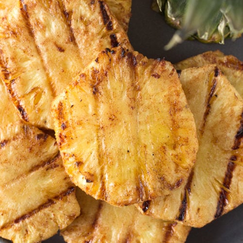 Top down shot of eight grilled pineapple slices on a gray plate with pineapple leaves in the upper right corner.