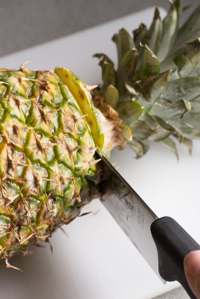 A sharp knife cutting the top off a pineapple on a white cutting board.