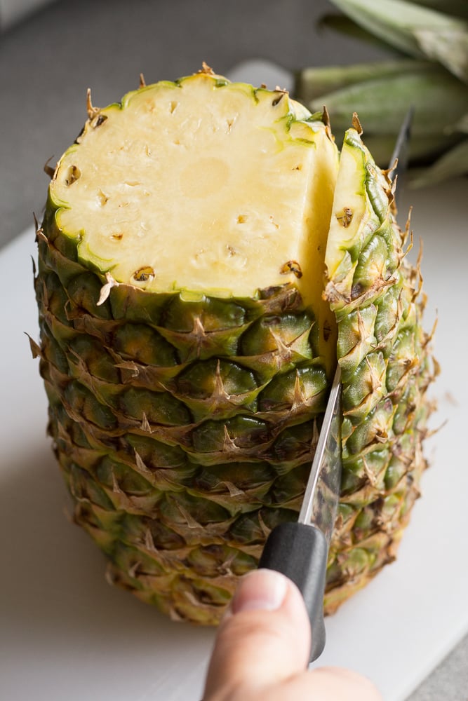 A large sharp knife cutting the skin off the sides of a pineapple on a white cutting board.