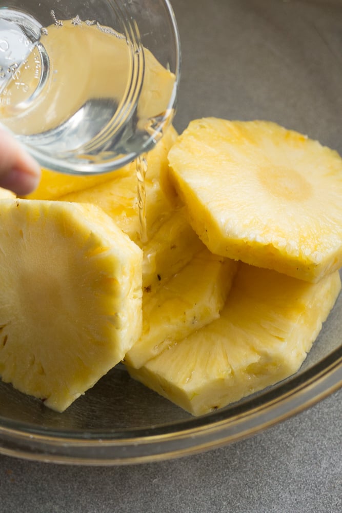 Pouring coconut oil out of a small clear bowl over pineapple slices in a large clear bowl.
