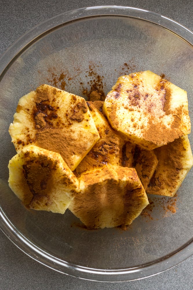 Slices of pineapple in a large clear bowl with cinnamon sprinkled on top.