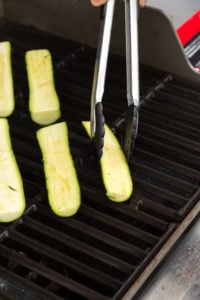 a pair of tongs placing halved zucchini on a grill