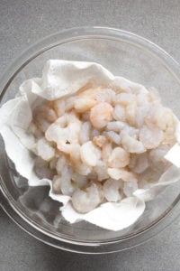 Top down shot of raw shrimp in a bowl with paper towels on a gray countertop.