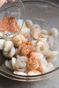 Sprinkling seasoning blend on raw shrimp in a large clear bowl.