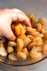 A hand rubbing seasoning blend onto raw shrimp in a large clear bowl.