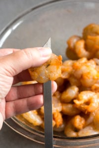 A hand placing a raw shrimp on a metal skewer.