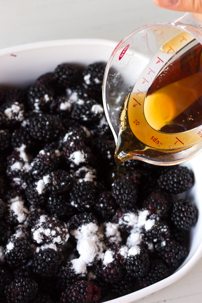 maple syrup being poured over blackberries in a baking dish.