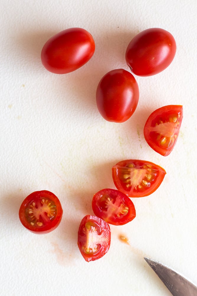 Cherry tomatoes being cut into quarters on a white cutting board.