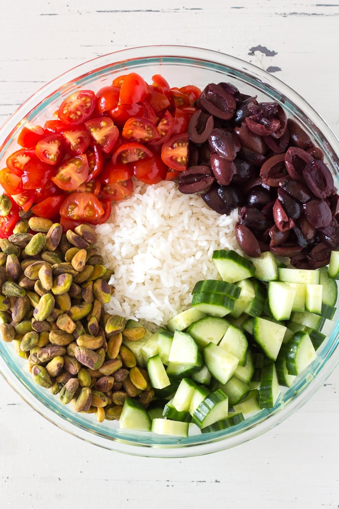 Top down shot of white rice, cut tomatoes, cut olives, pistachios, and cut cucumbers in a large bowl.