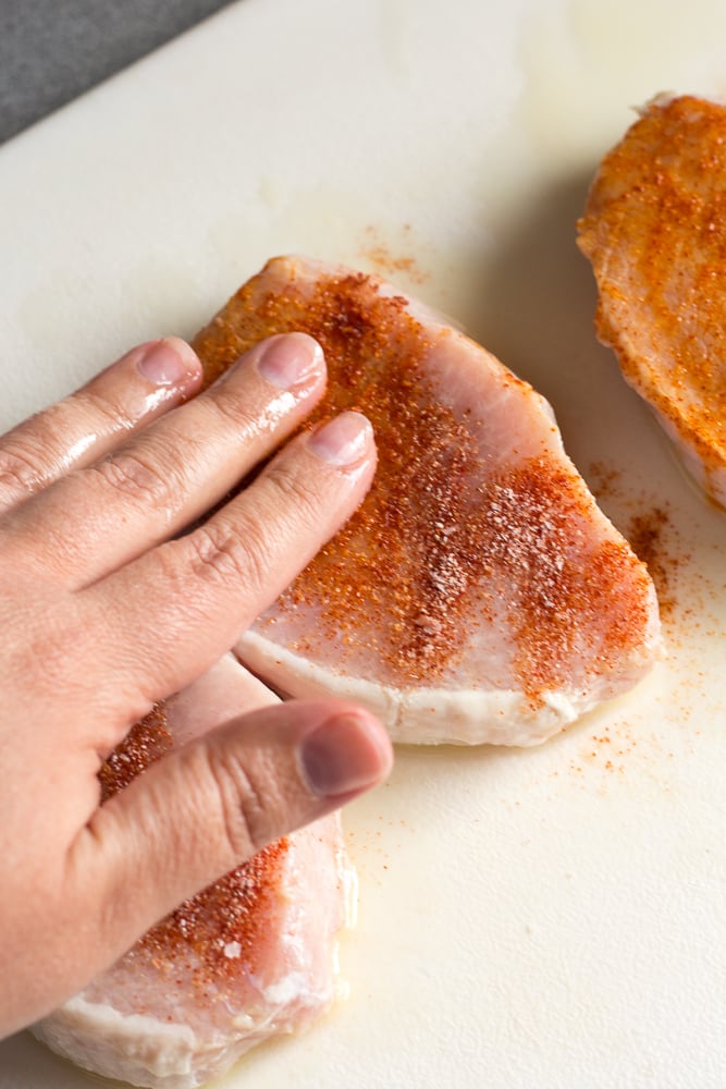 A hand rubbing seasoning onto raw boneless pork chops on a white cutting board.