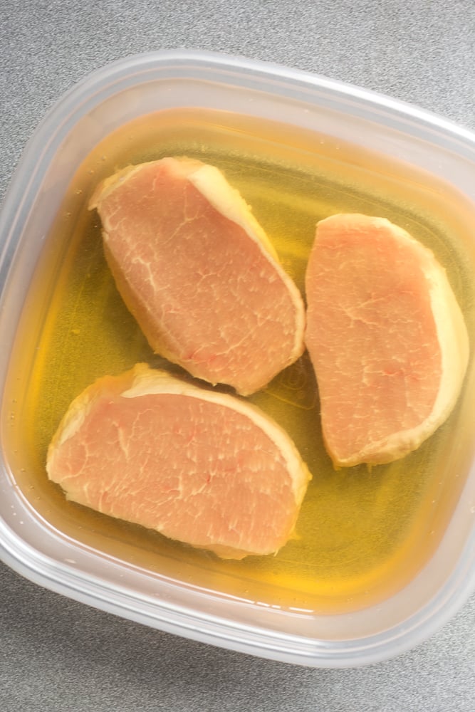 Three boneless pork chops in an apple juice brine solution all in a large plastic container.