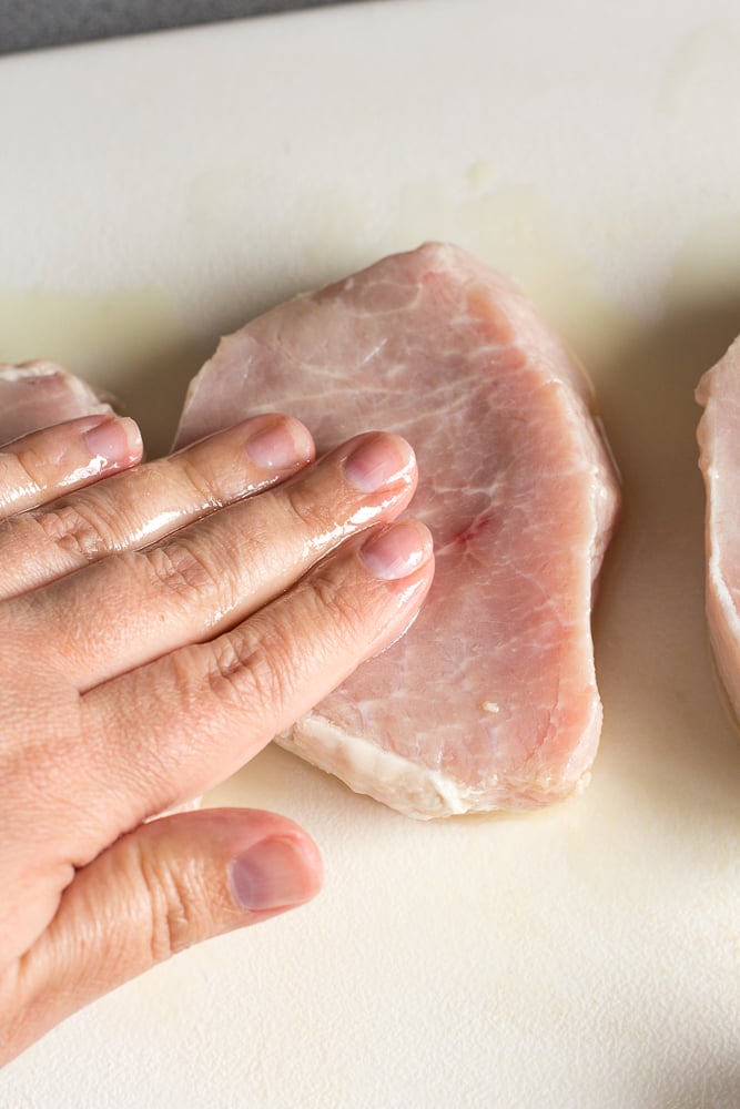 A hand rubbing oil on raw boneless pork chops on a white cutting board.