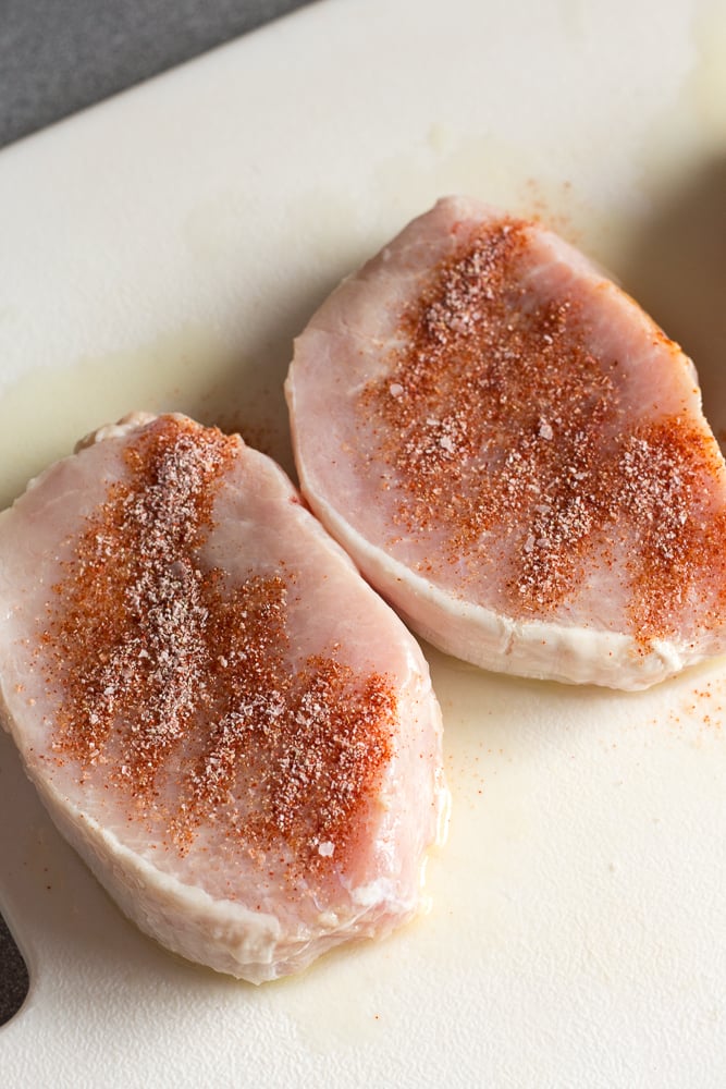 Seasoning sprinkled on raw boneless pork chops on a white cutting board.